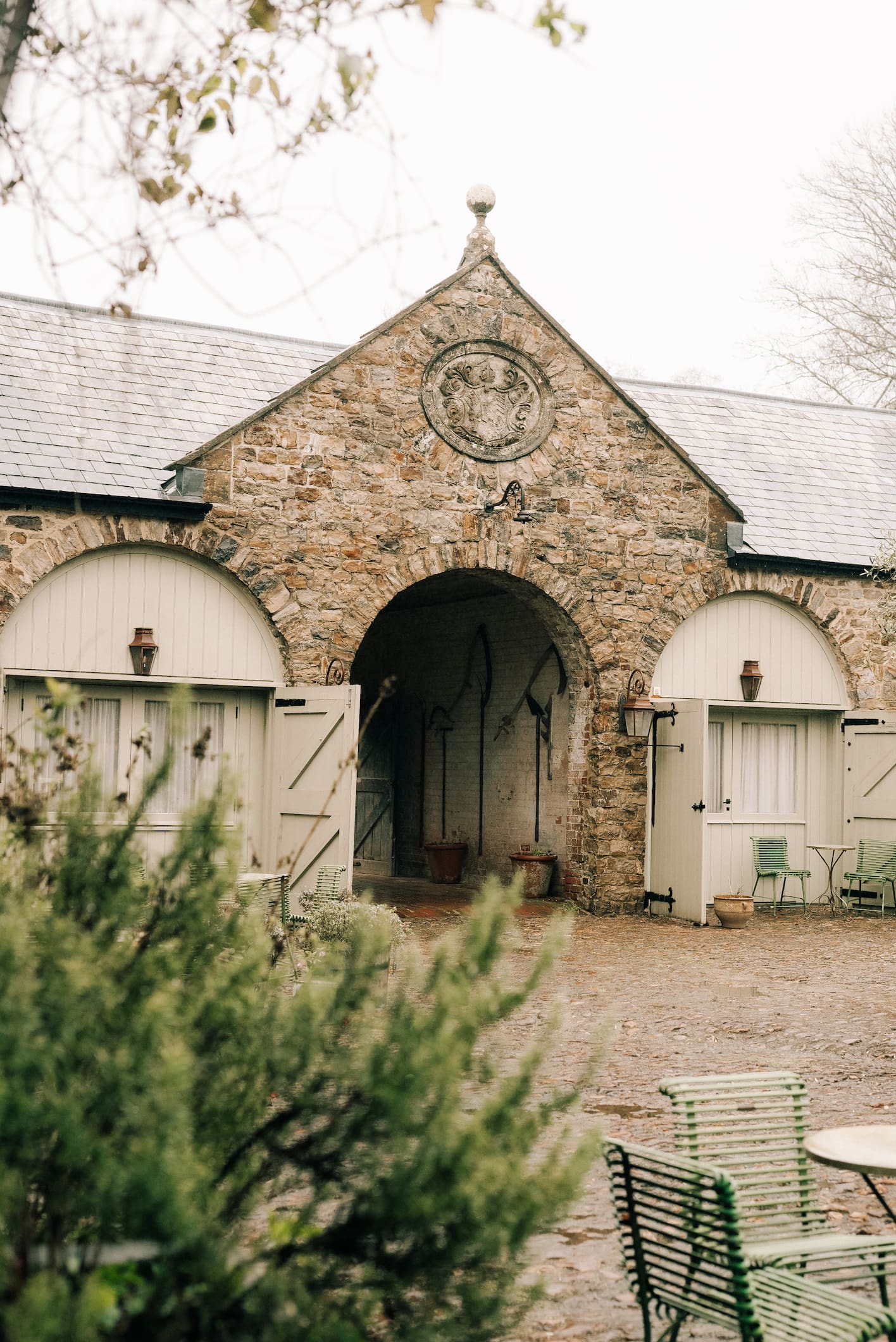 The stone stable courtyard at The Pig at Combe, with arched cream doors and green metal chairs