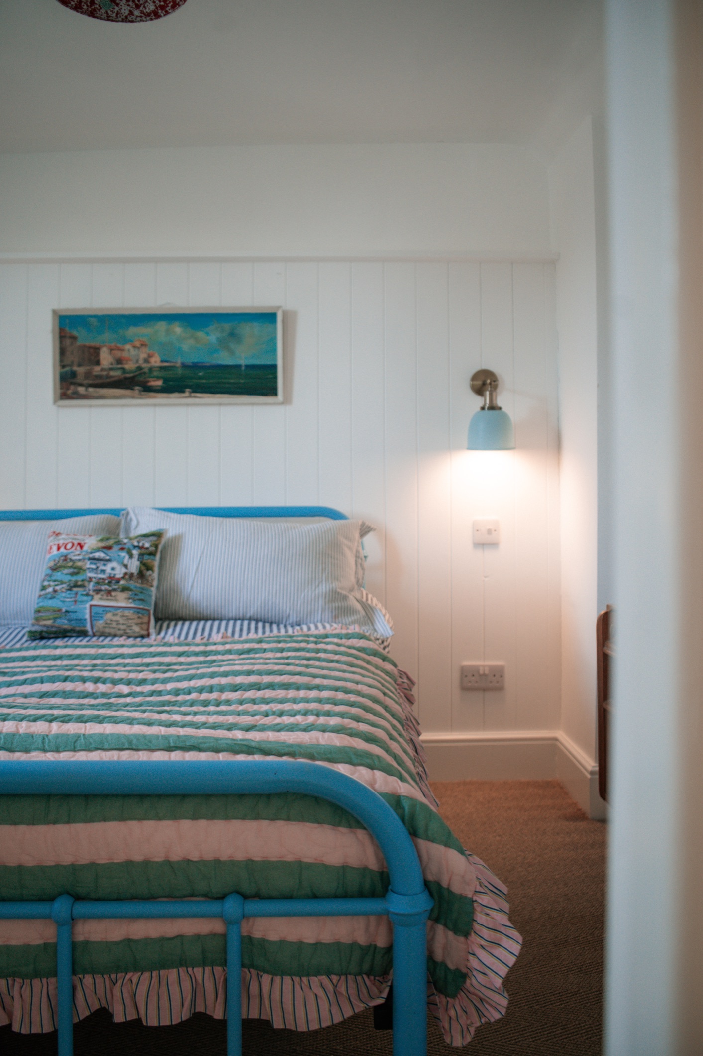 A blue iron bed with a pink and green striped quilt under a harbour painting at The Nook
