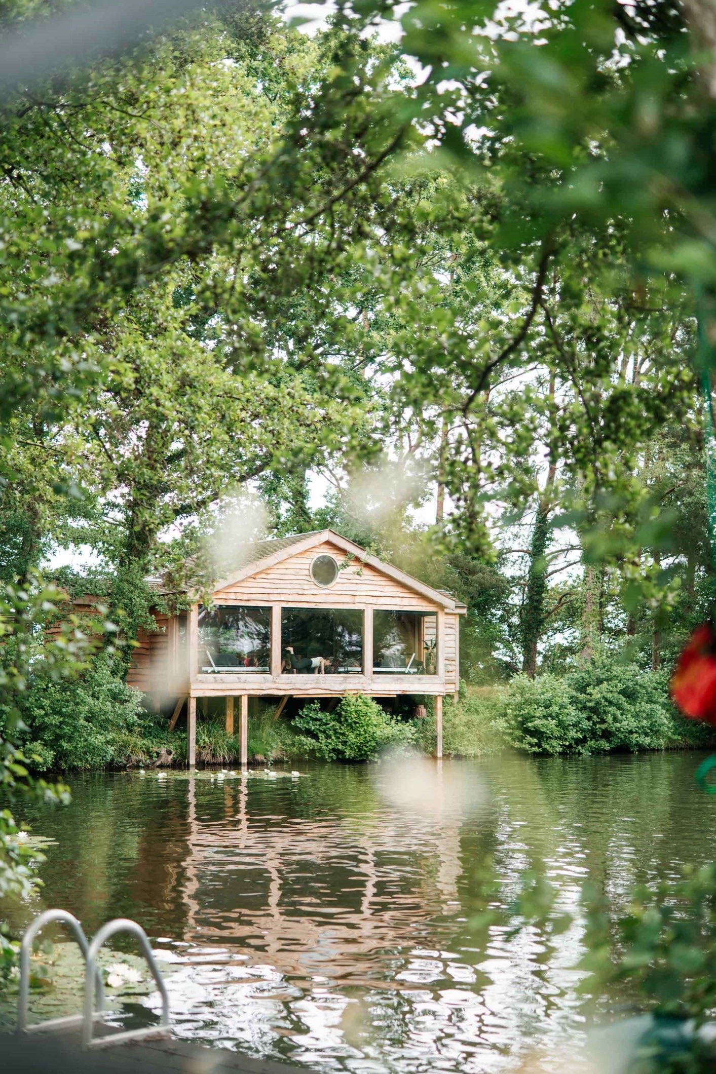 The timber cabin at Wild-Wood on stilts above a lake, seen through summer trees